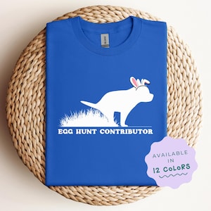 May include: A royal blue t-shirt with a white silhouette of a dog wearing bunny ears and the text "Egg Hunt Contributor". The text "Available in 12 Colors" is in a purple circle.