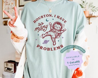 May include: A light blue t-shirt with the text "Houston, I have so many problems" and a cartoon cat astronaut. The text "Comfort Colors" is in a smaller font below the astronaut. The text "Available in 7 Colors" is in a purple box.