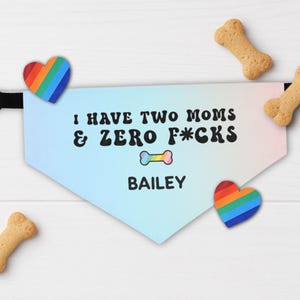 May include: A dog bandana with the text "I HAVE TWO MOMS & ZERO F*CKS" in black, with a rainbow bone graphic and the name "BAILEY". The bandana is light blue with a gradient effect and has two rainbow heart-shaped decorations. Four dog biscuits are scattered around.