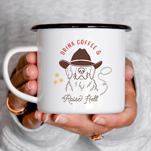 May include: White enamel mug with a black rim and handle. The mug features a brown illustration of a dog wearing a cowboy hat, with the words "DRINK COFFEE & Raise Hell" in brown and gold lettering. The mug is held in a person's hands.