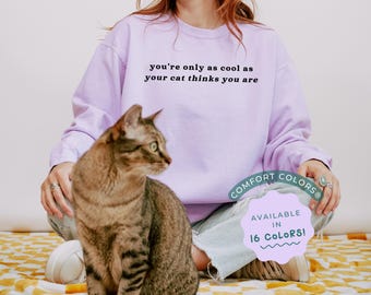 May include: A lavender long-sleeved sweatshirt with the text "you're only as cool as your cat thinks you are" printed on the front. A tabby cat is sitting next to the person wearing the sweatshirt. The sweatshirt is available in 16 colors.
