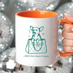 May include: White ceramic mug with an orange interior and handle. The mug features a teal illustration of a chihuahua in a handbag, with the text "...that's how much it hurts." The mug is set against a background of silver disco balls.