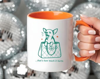 May include: White ceramic mug with an orange interior and handle. The mug features a teal illustration of a chihuahua in a handbag, with the text "...that's how much it hurts." The mug is set against a background of silver disco balls.