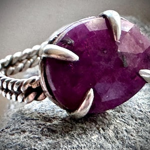 May include: A silver ring featuring a large, oval-shaped, deep magenta gemstone with a faceted surface. The stone is secured by four silver prongs. The ring band has a distinct twisted, rope-like texture crafted from polished silver metal.