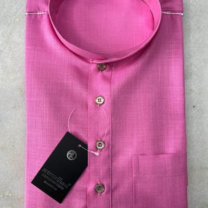 Puede incluir: Una camisa rosa brillante doblada con cuello mandarín y un bolsillo. La camisa tiene botones dorados y una etiqueta negra con texto blanco. La etiqueta dice "MATCH LINE AUTHENTIC WEAR".