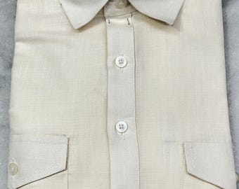 May include: Cream-colored button-down shirt with a classic collar and two chest pockets. The shirt is folded neatly, showcasing the buttoned front and the texture of the fabric. The shirt has a tailored fit.