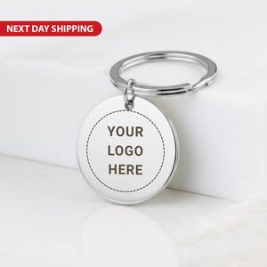 May include: A silver-colored round keychain tag with the text "YOUR LOGO HERE" in black. The tag is attached to a silver key ring. The background is white.
