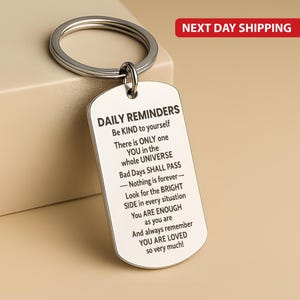 Könnte beinhalten: Ein silberfarbener Schlüsselanhänger mit einem rechteckigen Anhänger. Der Anhänger hat abgerundete Kanten und trägt den Text "DAILY REMINDERS" gefolgt von einer inspirierenden Botschaft. Der Schlüsselanhängerring ist ebenfalls silberfarben.