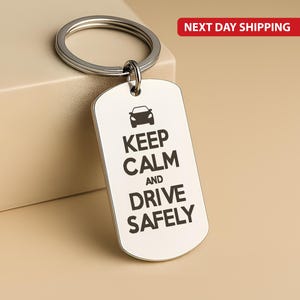 Puede incluir: Un llavero plateado con una etiqueta rectangular. La etiqueta presenta un icono de coche encima de las palabras "KEEP CALM AND DRIVE SAFELY" en negro y en negrita. El llavero tiene un anillo circular para sujetar las llaves.
