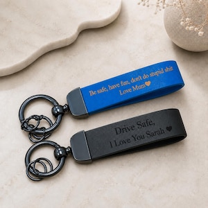 Op de afbeelding: Twee rechthoekige sleutelhangers met zwarte metalen ringen. De ene is blauw met de gegraveerde tekst: Be safe, have fun, don't do stupid shit Love Mum met een hartje. De andere is zwart met de gegraveerde tekst: Drive Safe, I Love You Sarah met een hartje.