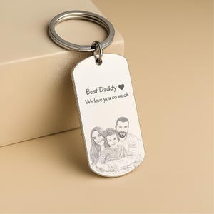 May include: A silver-toned metal dog tag keychain featuring a circular split ring. The surface is engraved with the text "Best Daddy" followed by a heart symbol and "We love you so much," along with a line art illustration of a family of three.