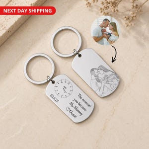 Op de afbeelding: Twee rechthoekige zilverkleurige metalen sleutelhangers met ringen. Eén hanger toont een gegraveerde lijntekening van een ouder met een baby. De andere hanger toont een gegraveerde klok, de datum 23.11.25 en de tekst The moment you became My Mummy Ocsar.