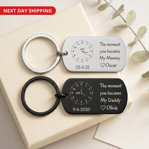 May include: Two rectangular keychains with a clock design. One is silver with the text "The moment you became My Mummy Oscar" and the date 23-11-25. The other is black with the text "The moment you became My Daddy Olivia" and the date 9-11-2020.