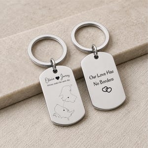 May include: Two silver-toned metal rectangular keychains with rounded corners and split rings. One keychain features the names Olivia and James, a heart, the text Alwasy under the same sky, and a map graphic. The other displays Our Love Has No Borders.