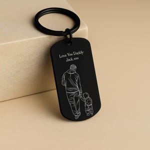May include: A matte black metal dog tag keychain featuring a white line art engraving of an adult holding a child's hand. The text "Love You Daddy Jack xxx" is engraved above the figures. The keychain includes a circular black metal ring.