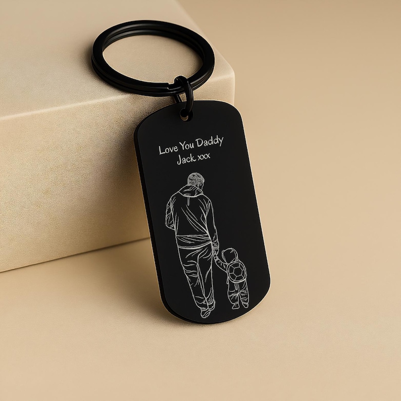 May include: A matte black metal dog tag keychain featuring a white line art engraving of an adult holding a child's hand. The text "Love You Daddy Jack xxx" is engraved above the figures. The keychain includes a circular black metal ring.