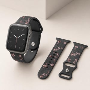 Op de afbeelding: Een zwarte smartwatch met een zwarte band met een roze bloemenpatroon. Twee extra zwarte horlogebandjes met hetzelfde bloemendesign worden ook getoond. De wijzerplaat van het horloge is zwart met witte en roze accenten.