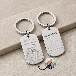 May include: Two silver-toned metal dog tag keychains with split rings. One tag features a line-art illustration of a couple, the names Chris and Laura, and the text The day we both said yes. The other tag displays an October 2025 calendar.