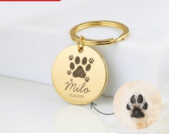 May include: A gold-colored, round keychain tag with a paw print design and the name "Milo" engraved. The tag also includes a date. The text "Custom Engrave your Pet Paw & Name" is at the bottom.