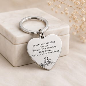 May include: A silver-coloured heart-shaped keyring featuring an engraved quote that reads: Sometimes amazing people forget how amazing they are... This is your reminder. Below the text is a small black line drawing of Winnie the Pooh and Piglet.