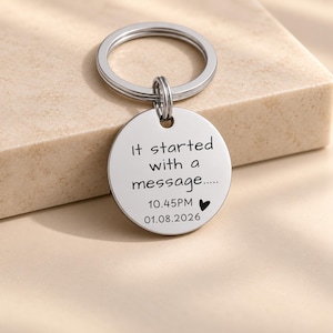 May include: A circular silver-coloured metal keyring featuring an engraved inscription that reads: It started with a message..... 10.45PM followed by a small black heart symbol and the date 01.08.2026. The pendant is attached to a standard split ring.