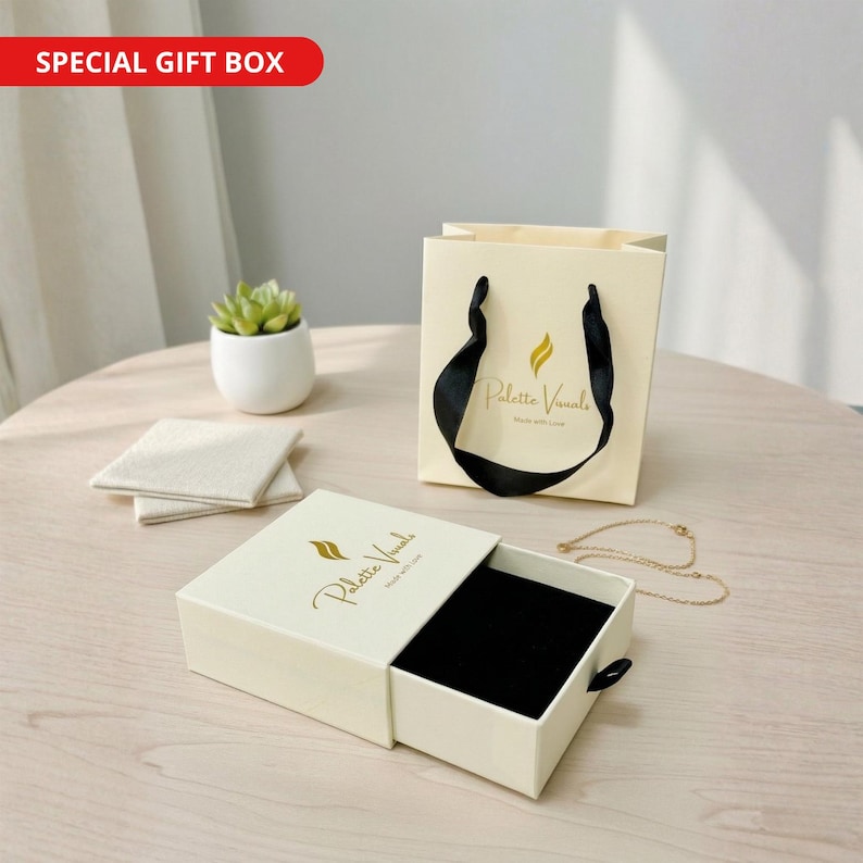 May include: A cream-colored sliding jewelry box and a matching cream-colored gift bag with black ribbon handles. Both items feature the gold text Palette Visuals. A thin gold chain necklace is placed next to the open box on the table.