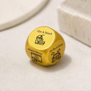 Pode incluir: Um dado de decisão de cor dourada com ícones e texto gravados em preto. Os lados visíveis exibem as frases Get A Snack com um ícone de bebida e sanduíche, Watch Movie com um ícone de filme e Go For A Drive com um ícone de carro.