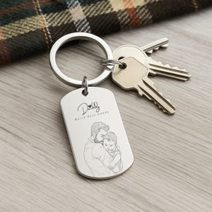 May include: A silver-toned metal dog tag keychain featuring an engraved line drawing of an adult holding a child. The text "Daddy" with a small heart icon and the phrase "World Best Daddy" are engraved above the illustration. Two metal keys are attached.