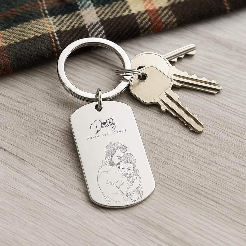 May include: A silver-toned metal dog tag keychain featuring an engraved line drawing of an adult holding a child. The text "Daddy" with a small heart icon and the phrase "World Best Daddy" are engraved above the illustration. Two metal keys are attached.