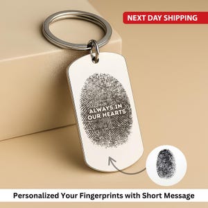 Op de afbeelding: Een zilverkleurige sleutelhanger met een rechthoekig label met een vingerafdrukontwerp en de tekst "ALWAYS IN OUR HEARTS". De sleutelhanger heeft een cirkelvormige ring voor het bevestigen van sleutels. Een kleine, cirkelvormige vingerafdrukgrafiek wordt ook getoond.