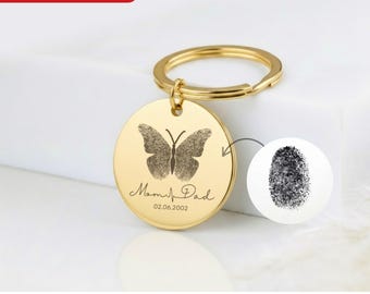 Könnte beinhalten: Ein goldfarbener Schlüsselanhänger mit einem runden Anhänger, der ein Schmetterlingsdesign aus Fingerabdrücken zeigt. Der Text "Mom & Dad" und ein Datum sind darunter eingraviert. Ein separates Fingerabdruckbild ist abgebildet. Der Text "Personalized Your Fingerprints in Butterfly with Your Name & Date" steht unten.