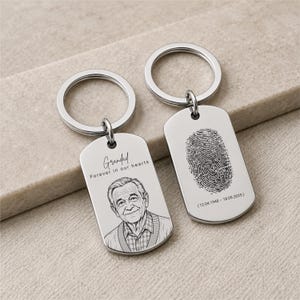 Puede incluir: Dos llaveros de metal plateado con forma de placa de identificación. Una placa presenta un retrato grabado de un hombre mayor, el texto "Grandad" y "Forever in our hearts". La otra muestra una huella dactilar grabada y "12.04.1948 - 18.09.2025".