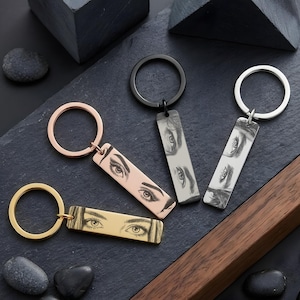 以下が含まれることがあります： 人間の目を刻印した4つの長方形の金属製キーホルダー。ゴールド、ローズゴールド、マットブラック、シルバーのキーホルダーが各1つずつあり、それぞれお揃いの円形リングが付いています。