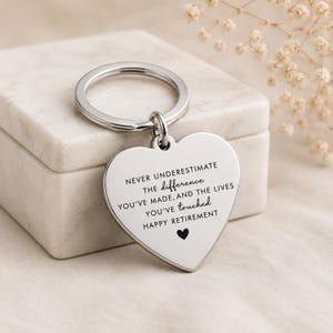 Pode incluir: Um porta-chaves prateado em forma de coração com um anel de metal. A superfície está gravada com o texto: NEVER UNDERESTIMATE THE difference YOU'VE MADE, AND THE LIVES YOU'VE touched HAPPY RETIREMENT, seguido de um pequeno símbolo de coração preto.