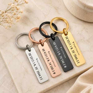 May include: Four rectangular metal keychains in silver, rose gold, black, and gold tones. Each features a split ring and is engraved with a location pin icon, a custom name, and geographic coordinates including 36°2317.1 N, 25°2544.3 E and 40°4121.3 N, 74°240.3 W.