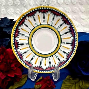 May include: A decorative ceramic plate with a white center, a yellow rim, and a detailed pattern in red, yellow, and blue. The plate is displayed on a clear stand, showcasing its intricate design. The plate's design is reminiscent of Italian pottery.
