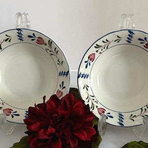 May include: Two white ceramic bowls with a blue rim and floral design. The bowls have a scalloped edge and are decorated with red and green floral patterns. The bowls are displayed on clear stands.