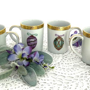 May include: Set of four white ceramic mugs with gold trim and painted Easter egg designs. Each mug features a different colored egg with a gold ribbon and bow. The eggs are purple, green, blue, and pink.