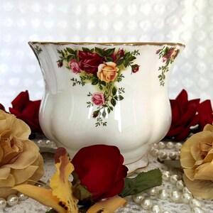 May include: A white ceramic planter with a scalloped rim and a gold trim. The planter is decorated with a floral pattern of red, pink, and yellow roses. The planter has a bulbous shape and sits on a small base.