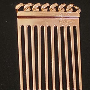 Puede incluir: Un peine dorado con un diseño de cadena en la parte superior. El peine tiene una fila de dientes y las palabras "VINTAGE" están grabadas en la parte posterior.
