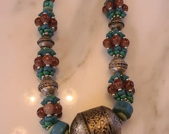 May include: A handcrafted necklace featuring a silver pendant with intricate designs and three dangling coin charms. The necklace incorporates turquoise, brown, and green beads, creating a unique and eye-catching accessory.