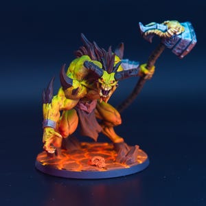 Puede incluir: Figura detallada de una criatura demoníaca con piel amarilla y naranja, cuernos negros y una expresión amenazante. Empuña un gran martillo y se alza sobre una base ardiente con una calavera. La figura está pintada con colores vibrantes.