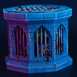 May include: A blue and purple miniature dungeon well with a barred window. The well is splattered with red paint, suggesting a horror theme. Inside the well, dice are visible. The well is designed for tabletop gaming.
