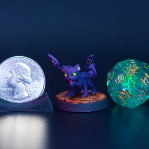 May include: A quarter coin next to a purple cat figurine with large ears and a green and gold d20 die. The cat figurine has a red tongue and is on a wooden base. The die is translucent with gold numbers.