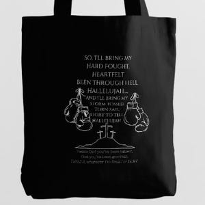 Könnte beinhalten: Schwarze Tragetasche mit weißem Text und Illustrationen. Das Design umfasst Boxhandschuhe, Kreuze und die Worte "SO, I'LL BRING MY HARD FOUGHT, HEARTFELT BEEN THROUGH HELL HALLELUJAH..." und andere Phrasen.