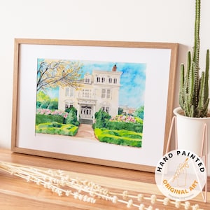 May include: A framed watercolor painting of a white house with a blue sky background. The painting is in a light brown wooden frame. The painting is labeled "Hand Painted Original Art".