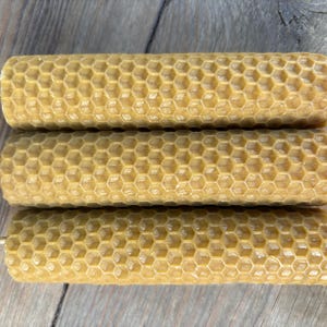 Puede incluir: Tres velas de cera de abeja natural con un patrón de panal. Las velas son de color marrón claro y están dispuestas en una pila.