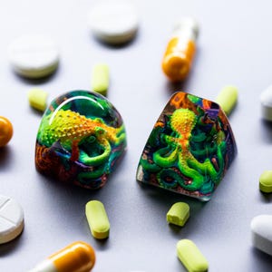 May include: Two resin keycaps featuring a colorful octopus with green, yellow, and orange tentacles. The keycaps are on a gray surface with various pills and capsules.