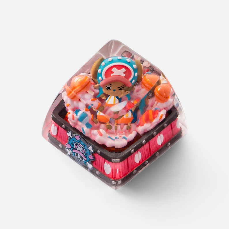 May include: A custom keycap featuring a cartoon character with a blue and white hat, orange accents, and a pink and white dessert-themed base. The keycap has a clear, transparent resin exterior and a red and black base with a skull and crossbones design.