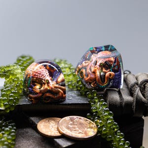 May include: Two resin keycaps featuring an octopus with iridescent purple, blue, and green accents. The octopus is depicted in a realistic style with a brown and orange color scheme. The keycaps are surrounded by green beads and copper-colored coins.